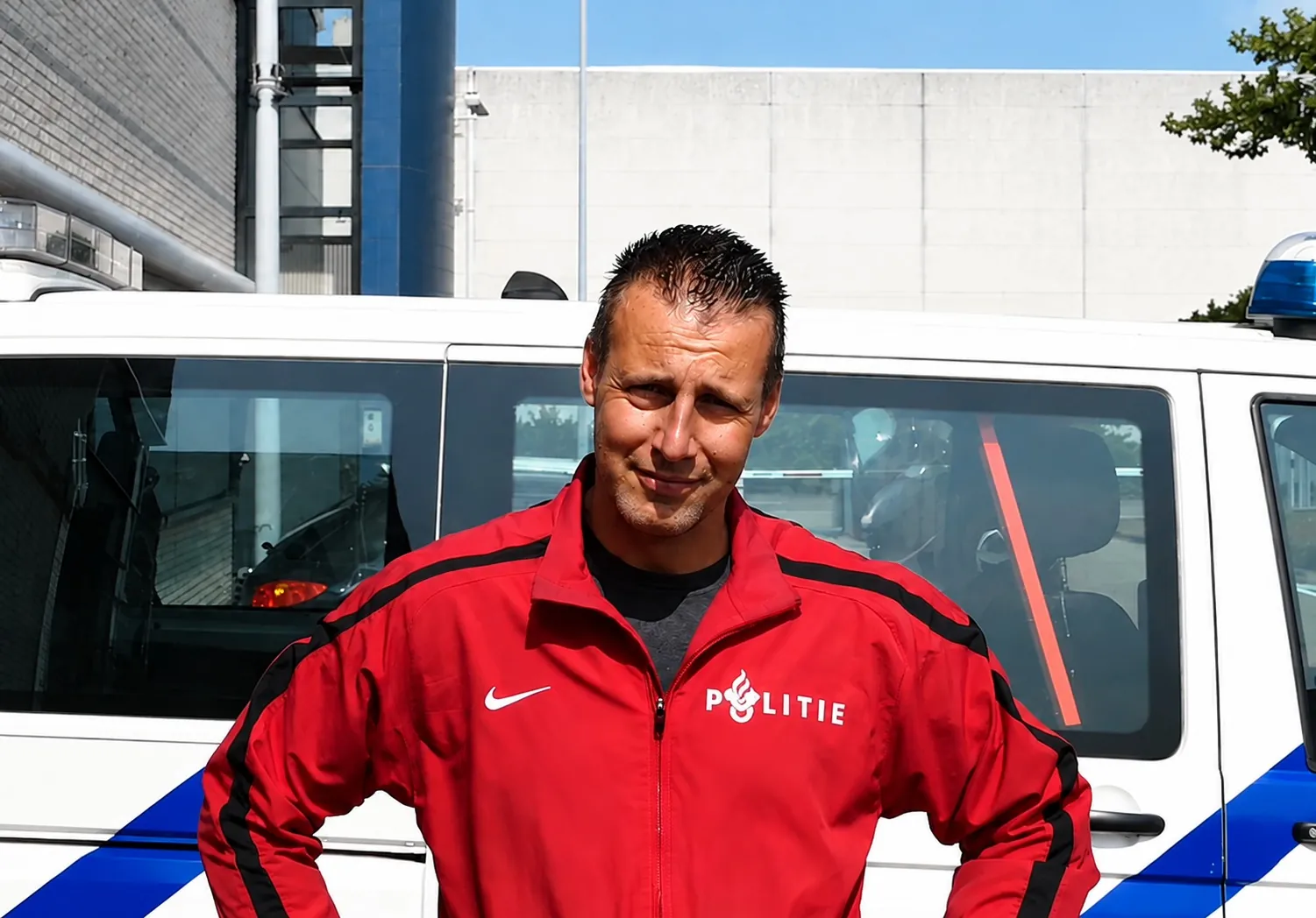 Maikel Menkhorst in een rood trainingsjack van de politie, voor een politiebus