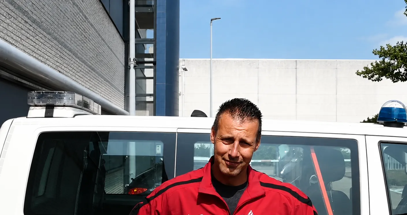 Maikel Menkhorst in een rood trainingsjack van de politie, voor een politiebus